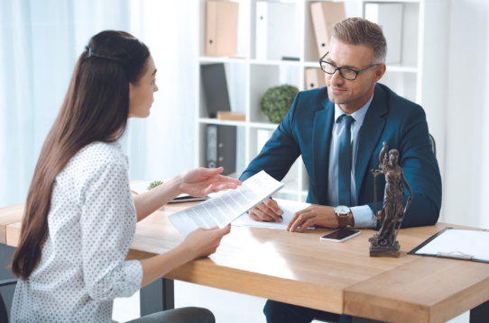A woman holding documents speaks with a lawyer in a blue suit at a desk, with legal figurines and paperwork visible, representing legal consultation