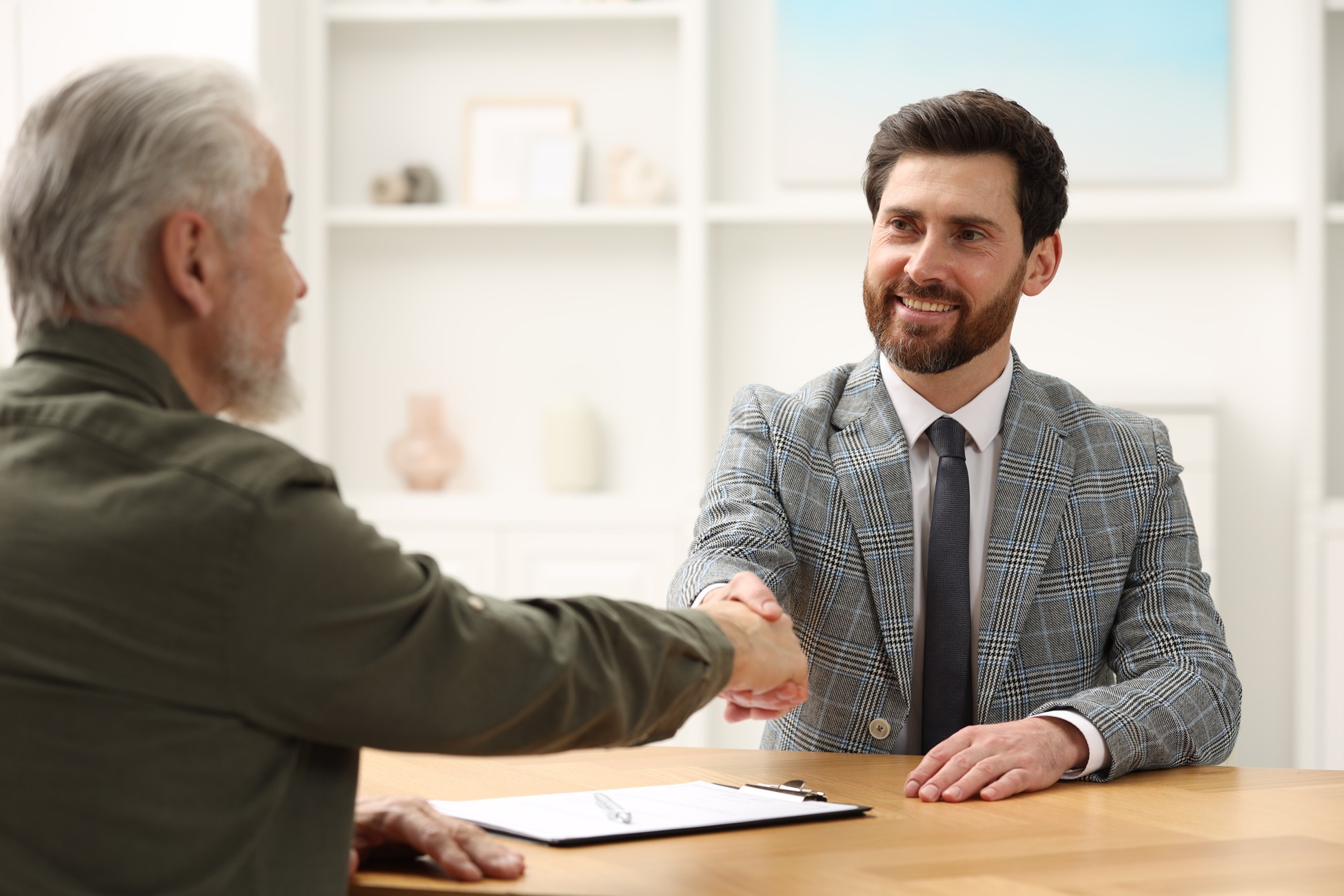 Senior man shaking hands with an attorney across a desk during a consultation, with a clipboard and pen in front of them.