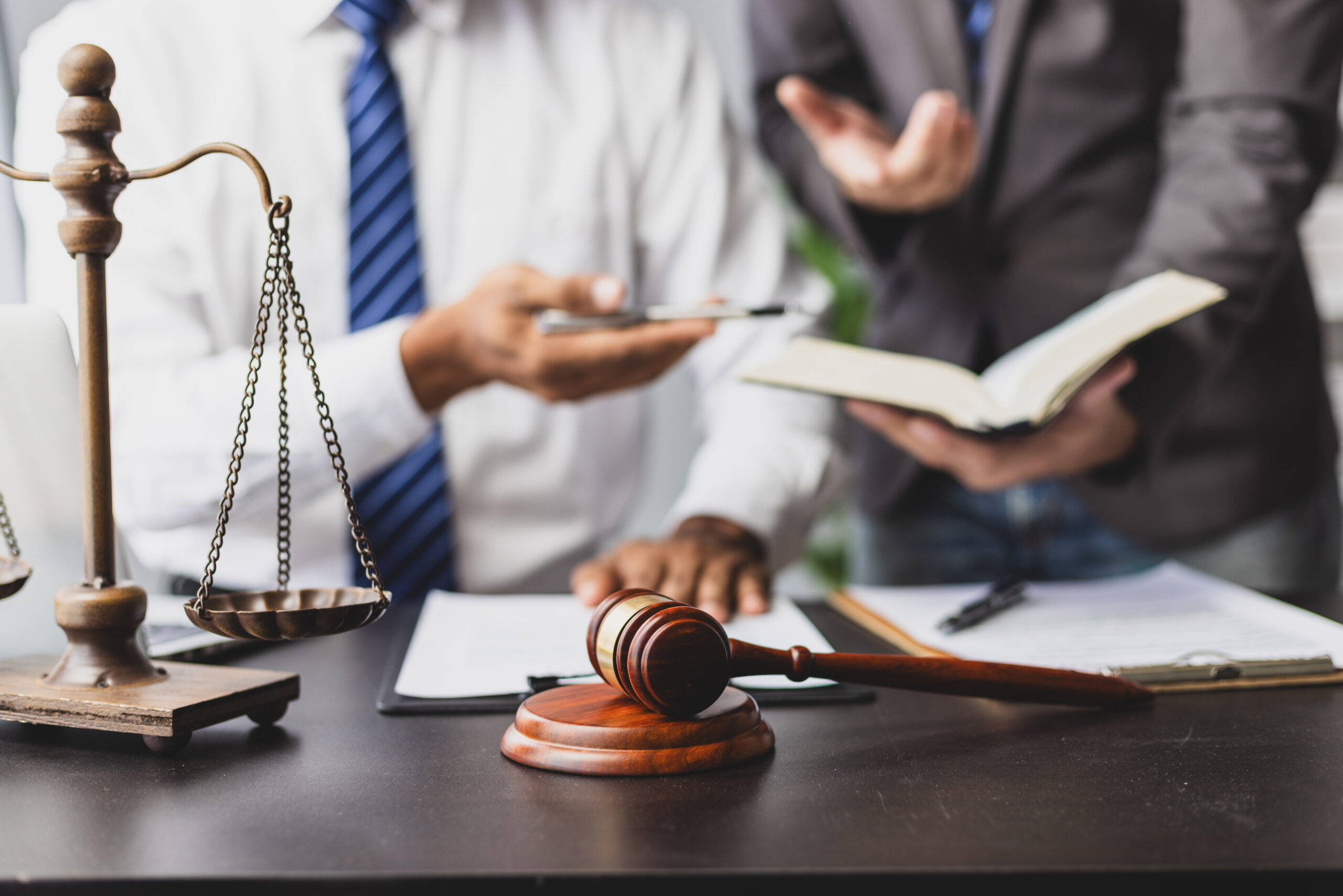 Close-up of a gavel and brass scales of justice on a desk, with two professionals in business attire discussing legal documents in the background.