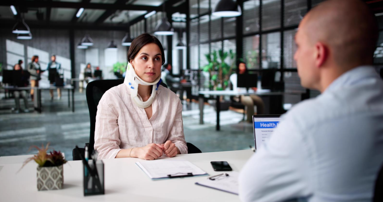 A woman wearing a neck brace sits at a desk in a modern office, listening to a man across from her during a meeting, with paperwork and a laptop on the table.