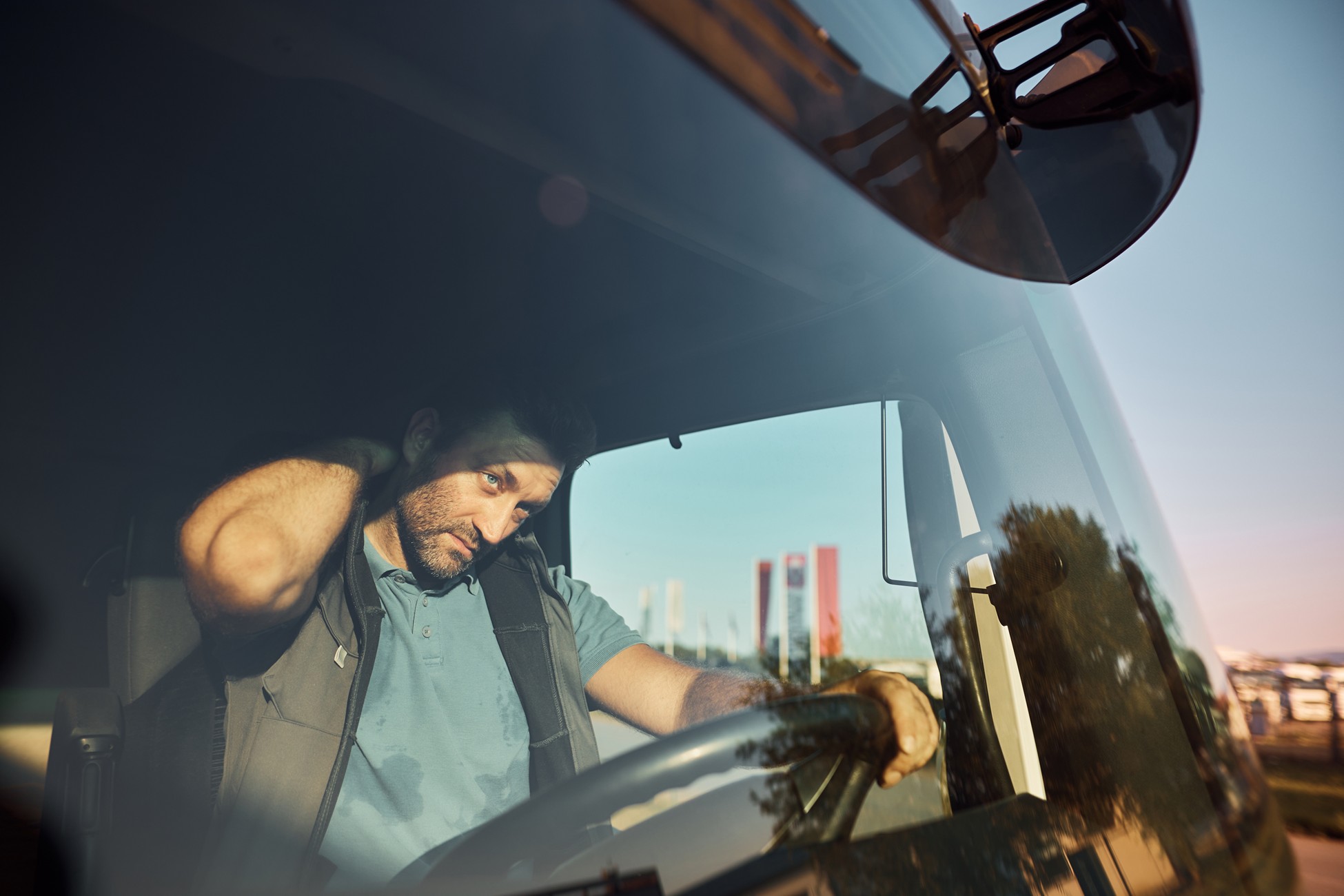 Truck driver inside his cab holding the back of his neck with a pained expression while seated at the steering wheel.