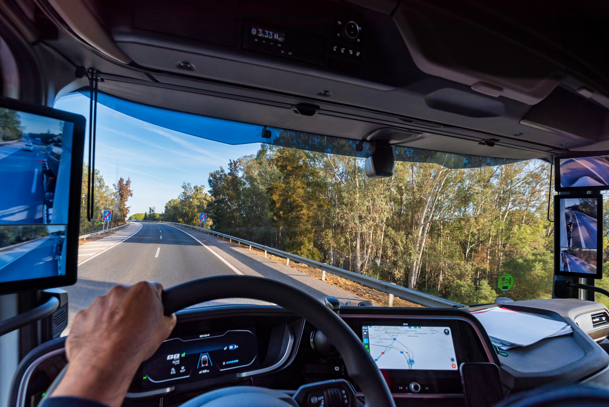 Commercial truck driver operating a vehicle with advanced digital dashboard displaying navigation and monitoring systems, with highway road and forest landscape visible through windshield