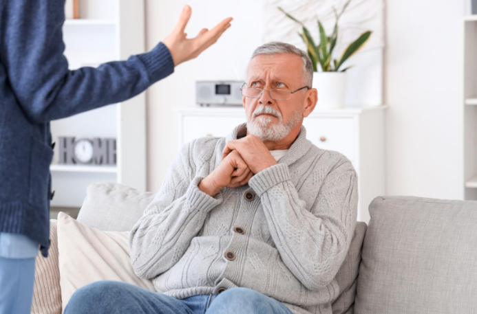 An elderly man sitting on a couch looking concerned and defensive as he faces a standing person gesturing with an outstretched hand, suggesting a tense or confrontational situation in a home setting.