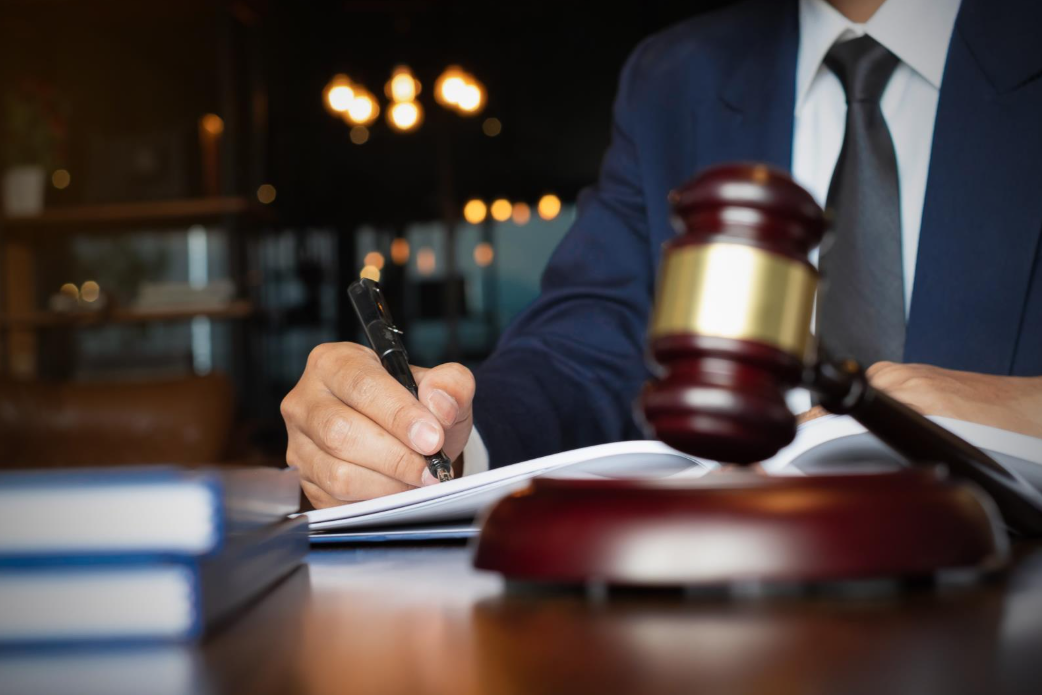 A close-up of a suited professional writing on documents at a desk, with a judge’s gavel in the foreground and law books blurred in the background.