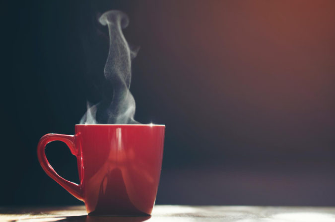 A red ceramic coffee mug with steam rising from it, placed on a wooden surface and illuminated by warm light.