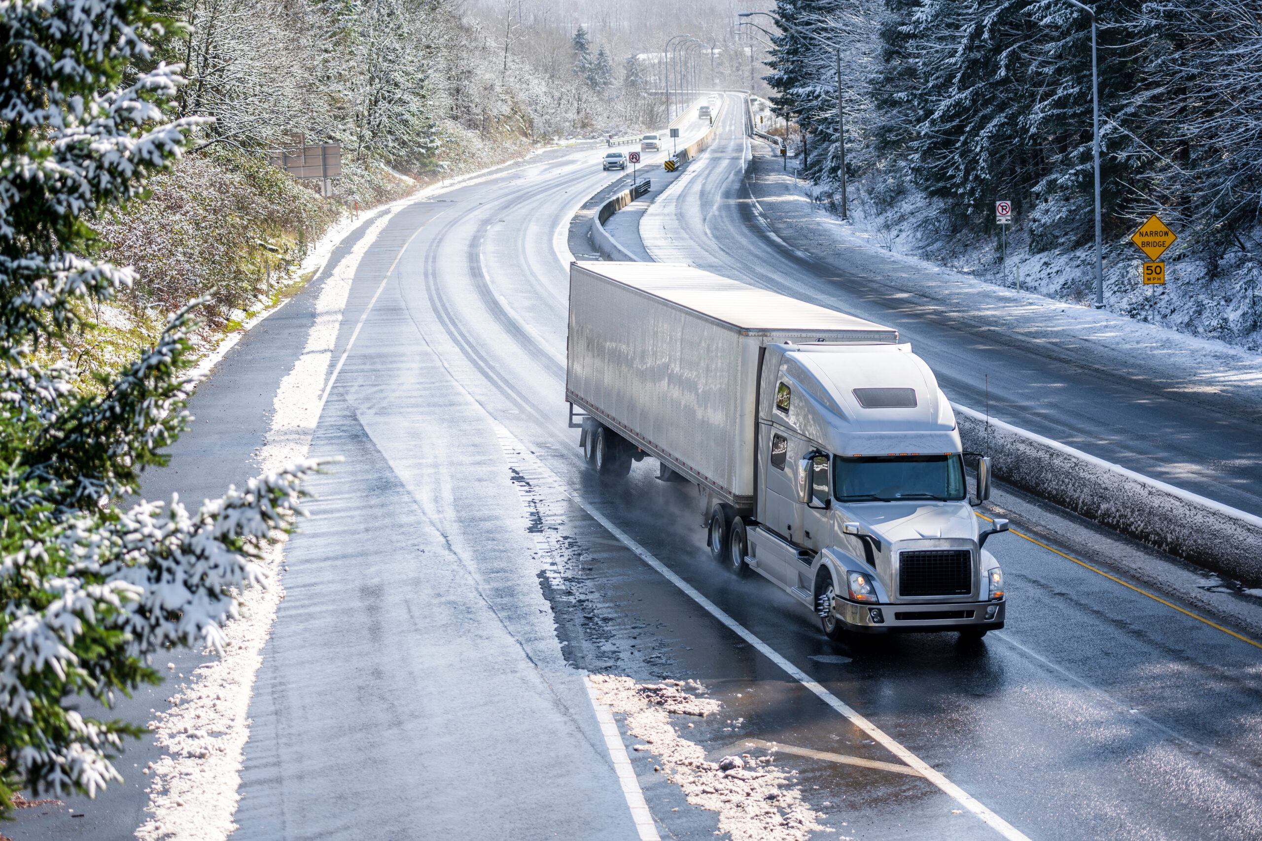 A semi-truck drives along a wet, icy highway surrounded by snow-covered trees and hills on a cold winter day.
