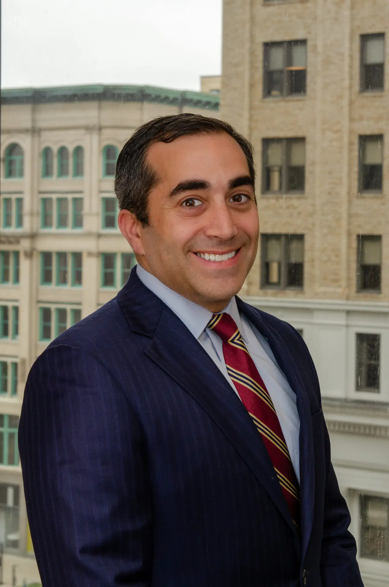 Buffalo attorney in navy pinstripe suit and striped tie smiling with downtown Buffalo buildings visible in background